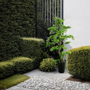 KĂĽnstlicher, detaillierter Bonsai mit schwarzem Topf in einem Aufstellbeispiel neben GebĂĽsch und Steinboden