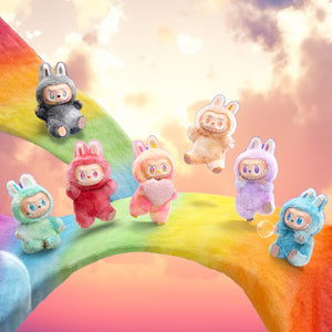 Sechs kleine PlĂĽschtiere in verschiedenen Farben auf einem Regenbogen mit Wolken im Hintergrund